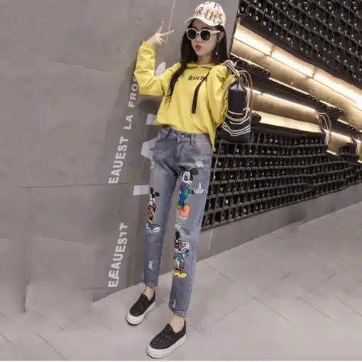 Noor naine seisab tänaval, kandes Miki-Hiirega naiste teksaseid, millel on värvilised tegelased, kollast pusa ja musti kingi. Taustal on betoonsein ja riiulid.