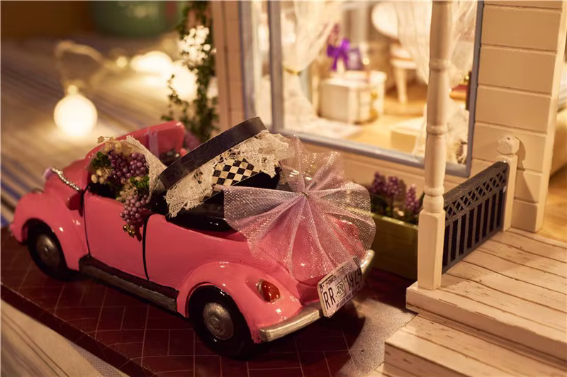 roosa miniatuurne auto lillede ja paeltega nukumaja ees