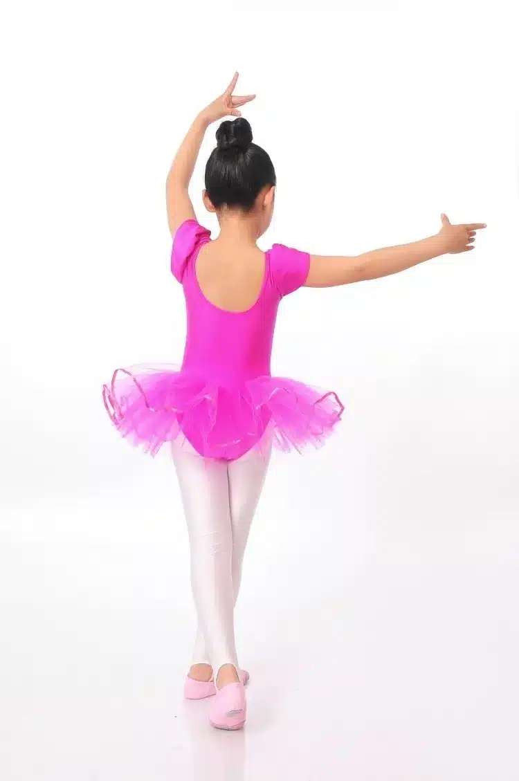 Tüdruk roosas baleriinikleidis, mis on erkroosa tutu seelikuga, seisab balleti poosis valgel taustal, seljas valged sukkpüksid ja roosad baleriinikingad.