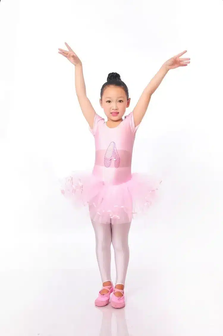 Roosas balletikleidis tüdruk, käed üles tõstetud, seisab valgel taustal. Kleit on kaunistatud õrnade tüllikihtidega, mis lisavad liikuvust ja elegantsi.