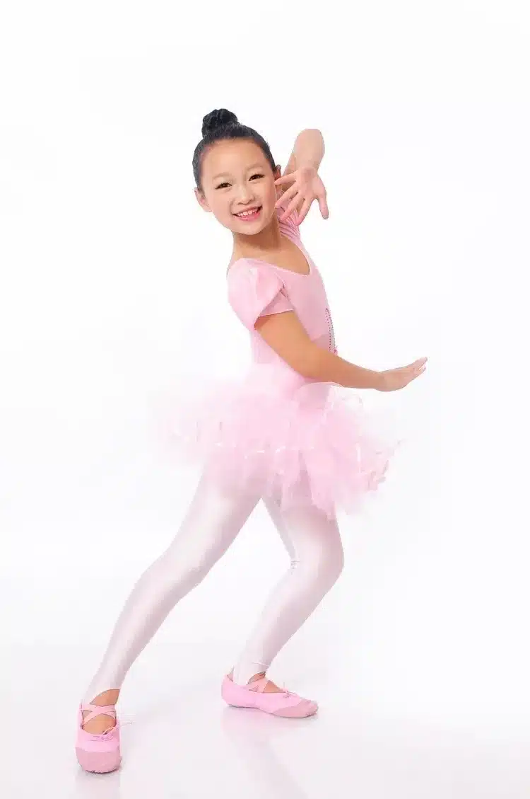 Tüdruk on riietatud imekaunisse balletikleidisse, millel on pehme roosa värv ja kohevad kihid. Taust on puhas ja valge, mis toob esile kleidi elegantsi ja disaini ilu. Lisaks on tüdrukul valged sukkpüksid ja roosad balletisussid.