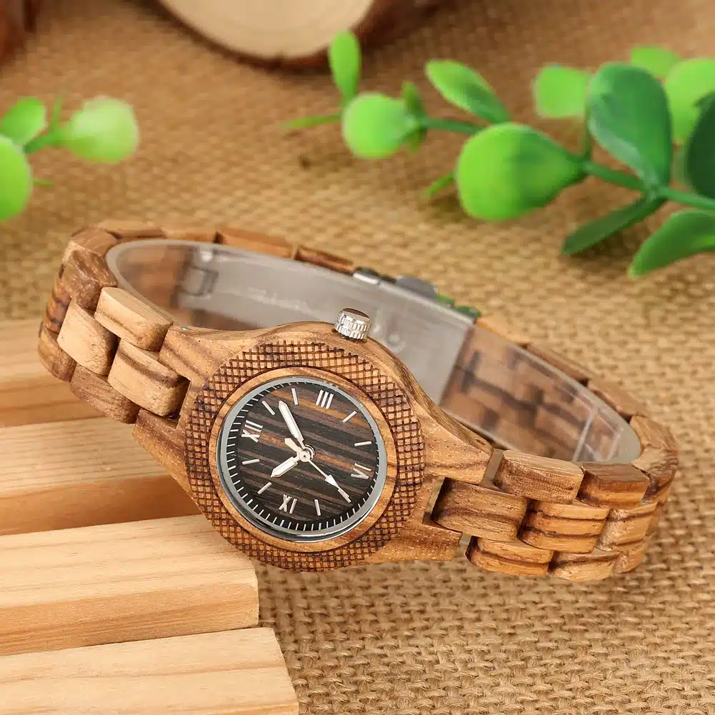 Stiilne puidust naiste käekell tumeda sihverplaadiga, rooma numbritega ja elegantsel heledast puidust rihmaga kangataustal, roheliste lehtede kõrval.