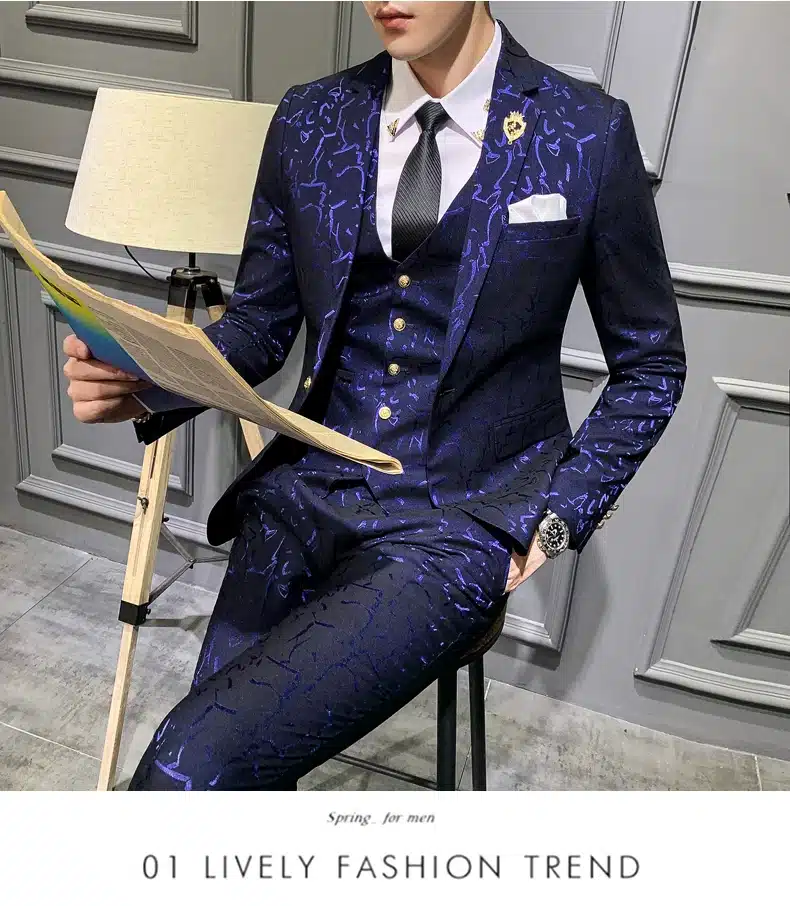 Mees istub toolil, kandes tumepunast mustrilist ülikonda. Taustal on hallid seinad ja lamp, mis lisavad stiilset atmosfääri.