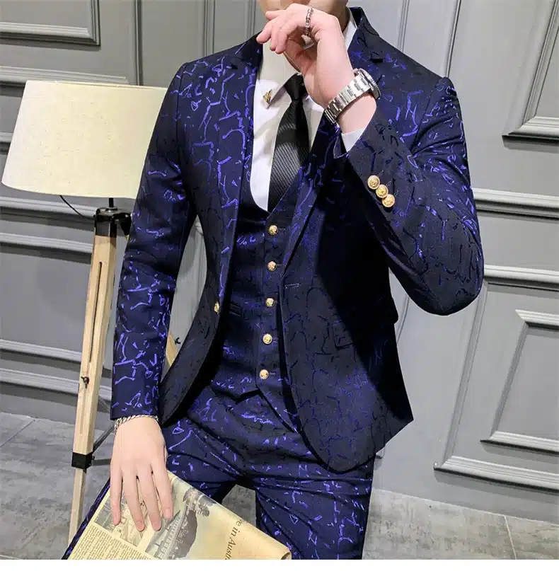 Mees kannab tumepunast mustrilist ülikonda valge särgi ja lipsuga, taustal hall paneelsein ja põrandal seisev lamp.