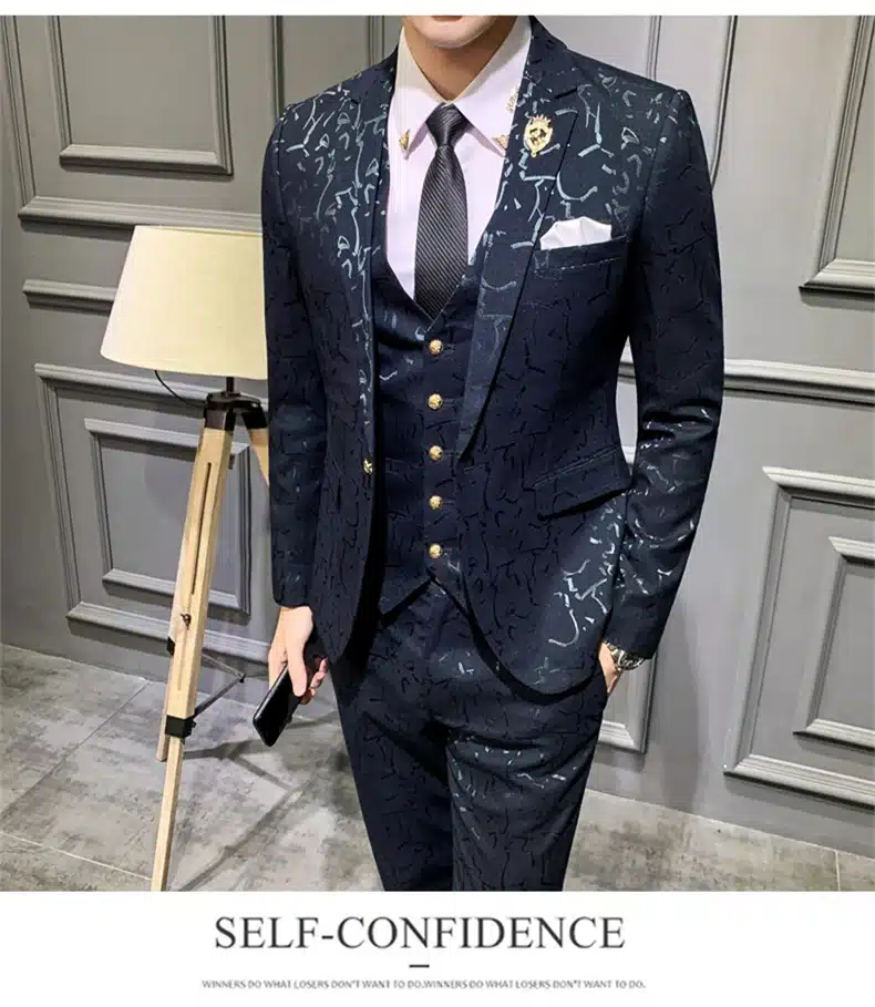 Elegantne mees, kes kannab tumepunast mustrilist ülikonda. Ülikond koosneb lipsust, vestist ja pintsakust, millel on detailirohke muster. Taustal on hele hall sein ja põrandal lamp, rõhutades ülikonna stiilsust.