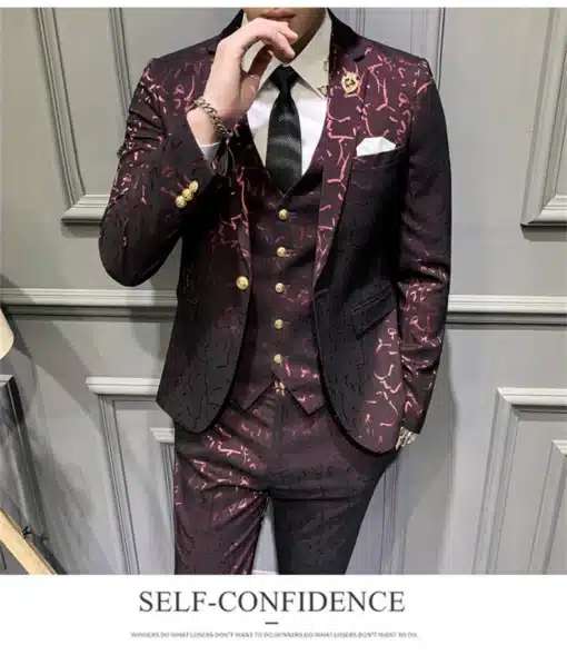 Stiilne tumepunane mustriline ülikond, kuldsete nööpidega pintsakul, valge särgi ja musta lipsuga halli paneelseina taustal.