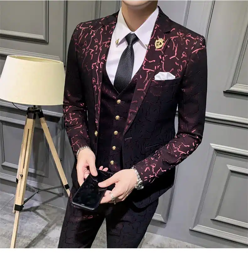 Tumepunane mustriline ülikond, mida kantakse halli paneelseinte taustal. Ülikond koosneb mustast lipsust, valgest särgist, kolmeosalisest tumepunasest vestist ja mustriga pintsakust ning pükstest. Pildil on käes ka nutitelefon.