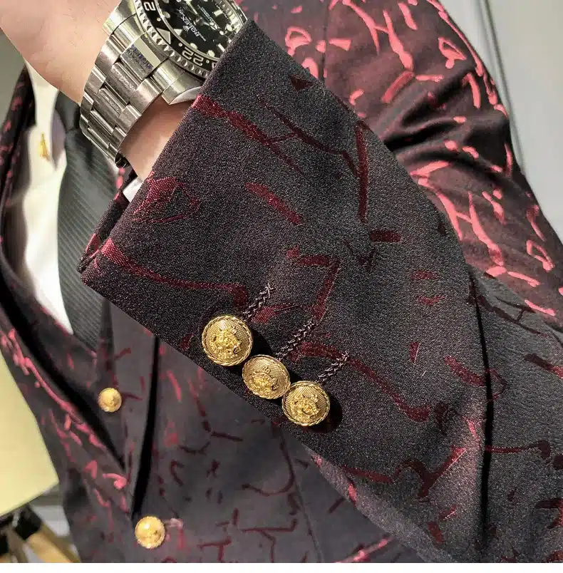 Elegantses keskkonnas demonstreeritakse tumepunast mustrilist ülikonda, millel on kuldsed nööbid ja keerukas muster. Ülikonda kantakse koos kella ja lipsuga, lisades üldisele stiilile luksust.