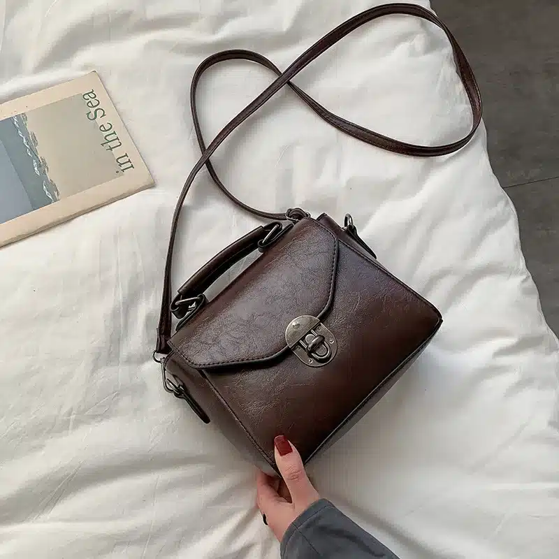 Elegantne, tumepruun väike crossbody kott sangaga, mida hoiab käsi, lamades valgel voodilinal raamatu kõrval.