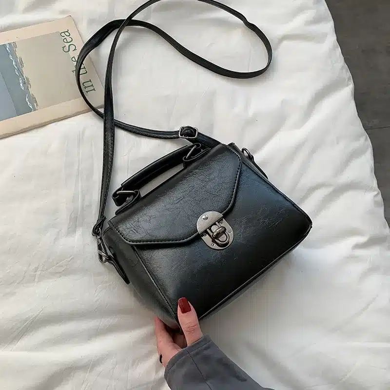 Ehtne must nahast väike crossbody kott sangaga, musta rihmaga, valgel voodil lebamas. Koti eesosas on metallist lukk.