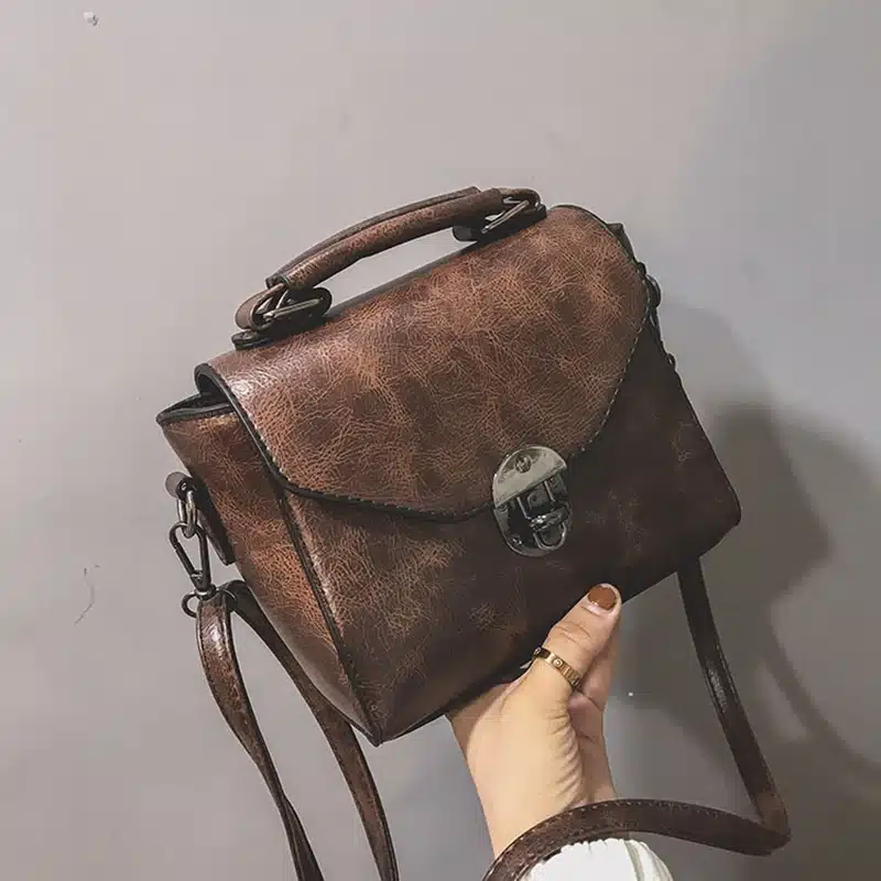 Käsitsi hoitud pruun väike crossbody kott sangaga, valmistatud kvaliteetsest nahast, millel on kaunis läikiv viimistlus ja hõbedane lukk, hallil taustal.