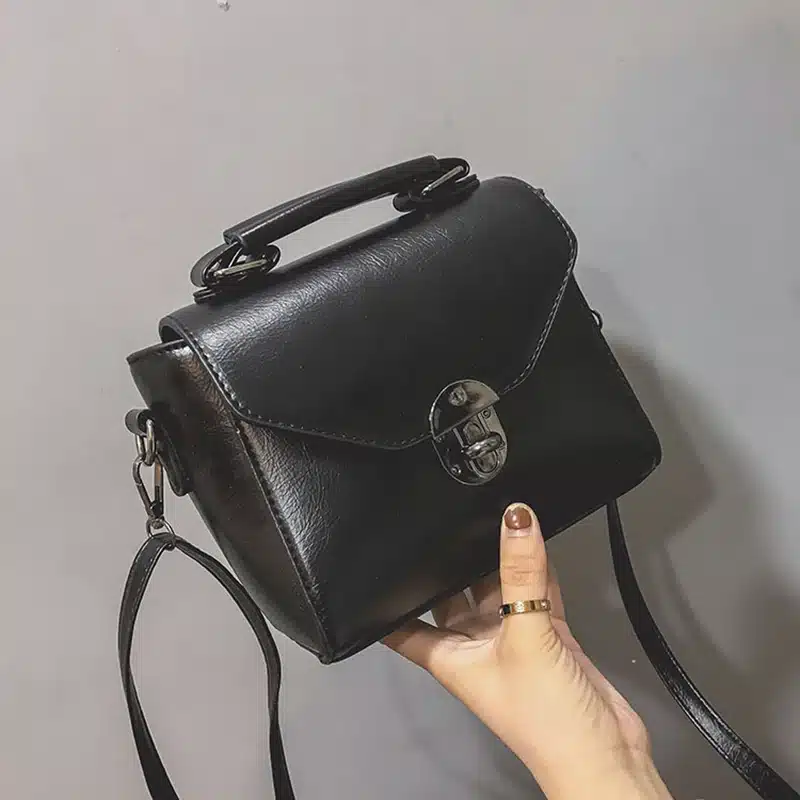 Must väike crossbody kott sangaga, mis on valmistatud sileda nahataolise materjaliga. Kotil on hõbedane lukk ja reguleeritav rihm. Taust on hall ja minimalistlik, esile tõstes kotti ja selle elegantset disaini.