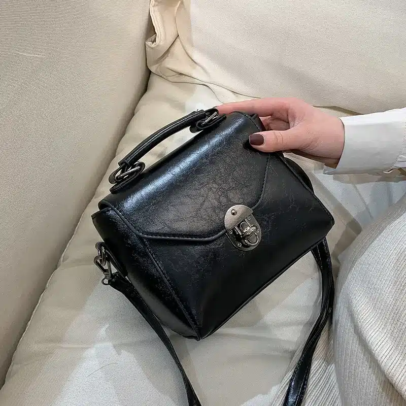 Väike must crossbody kott sangaga, valmistatud nahast, millel on metallist lukk esiküljel. Kott asub heledal tekstiilist taustal, käes hoiab seda naine, kellel on tume maniküür.