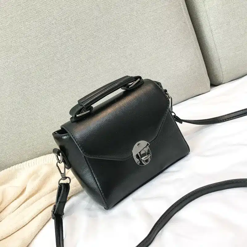 Elegantne must väike crossbody kott sangaga, mis on asetatud pehmele valgele kangale ning taustaks hele diivan.