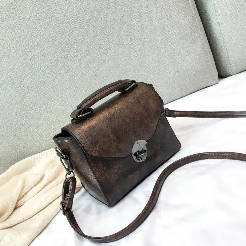 Stiilne pruun väike crossbody kott sangaga, mis on valmistatud nahast. Kott asub helehallil diivanil, valgel taustal, ja sellel on kroomitud lukk.