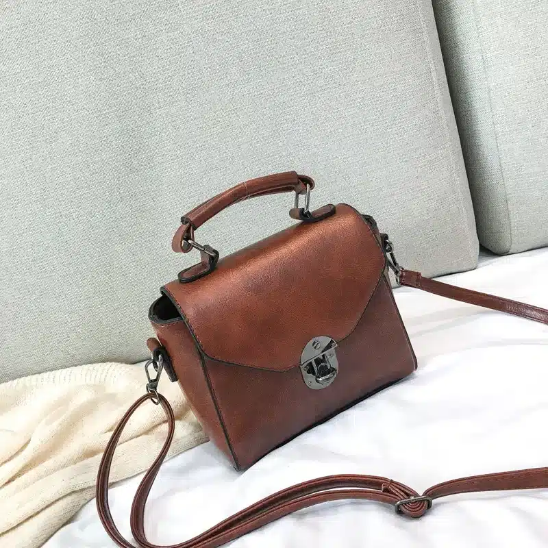 Pruun nahast väike crossbody kott sangaga, asetatud valgele voodilinadele, taustal hele diivan.