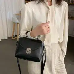 Naine kannab väikest musta crossbody kotti sangaga, millel on hõbedane lukk. Kotil on klassikaline disain ja see sobib elegantse beži pluusi taustaga hubases interjööris.