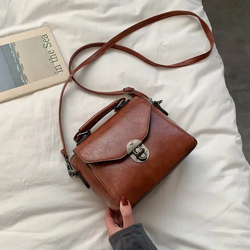 Pruun nahast väike crossbody kott sangaga, mis lebab valgel voodikattel koos avatud raamatu kõrval. Koti klamber ja sanga kinnitused on metallist, andes sellele stiilse ja klassikalise välimuse, samal ajal kui käsi hoiab sellest kinni.