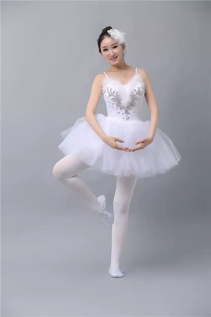 Naine poseerib klassikalises balletipoosis, seljas valge uhke balletikleit, mis on kaunistatud elegantsete hõbedaste detailidega. Kleit on kohev ja valmistatud tihedast tüllist, taust on helehall ja minimalistlik.