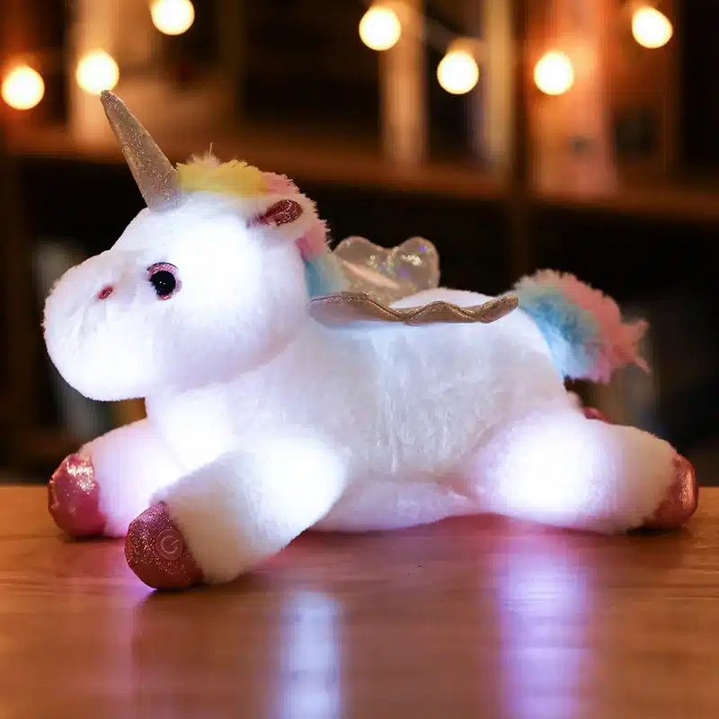 Led valgustusega pehme ükssarvik, valge ja vikerkaarevärviliste detailidega, lamab puidust laual, taustal hubane valgus.