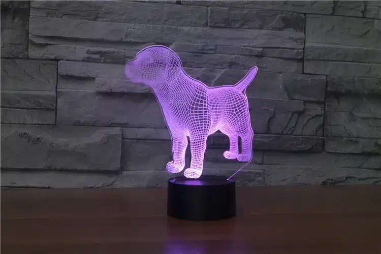 3D lamp 'Koer', mis kuvab lilla koera hologrammi kiviseina taustal, mustal alusel. Lamp pakub ainulaadset ja kaasaegset LED-valgustust ning sobib hästi igasse interjööri.