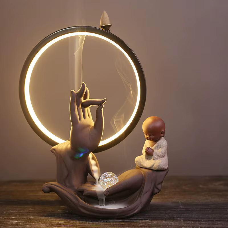 Viirukipõletaja kahe käega, kus keraamiline käsi hoiab suitsu asemel valgustatud ringi. Taustal on puitlaud, viirukipulk paistab mudeli kohal, täiendades rahuliku ja meditatiivse atmosfääri.