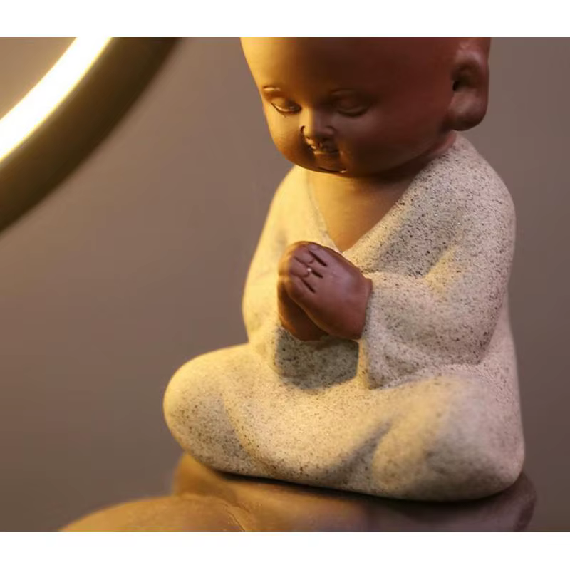Lähedalt pilt meditatsioonis poisisarnast kuju, millel on pruun savist pea ja heledast materjalist kehas, hoides oma käed palves poosis koos. Taustal on hämar valgustus, mis lisab rahuliku jaubaluure atmosfääri. See on osa tootest Viirukipõletaja kahe käega.