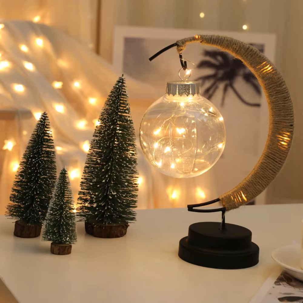 kuukujuline LED lauavalgusti, mille küljes ripub klaaskera, taustal väikesed kunstkuused