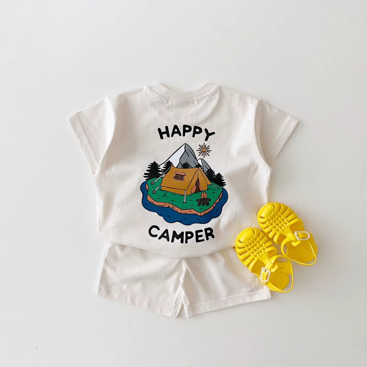 Beebi riietekomplekt 'Happy Camper', mis sisaldab heledat T-särki telkide ja mägede mustriga ning heledaid pükse, komplektis kollased sandaalid valgel taustal.