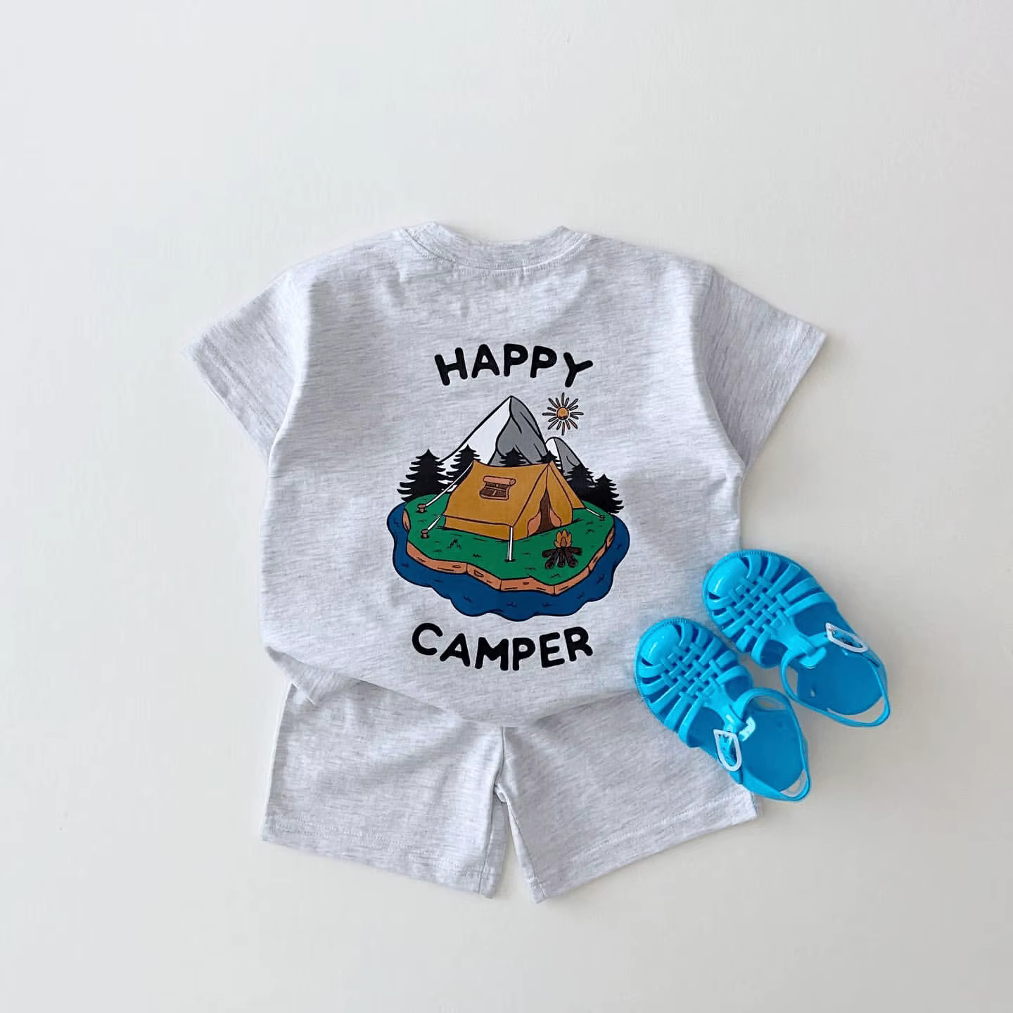 Komplekt 'Happy camper' koosneb hallist T-särgist ja lühikestest pükstest. T-särgi seljal on värviline pilt matkatelgist ja mägedest, ümbritsetud päikese ja puude motiividega. Kõrval on sinised lapse sandaalid valgel taustal.