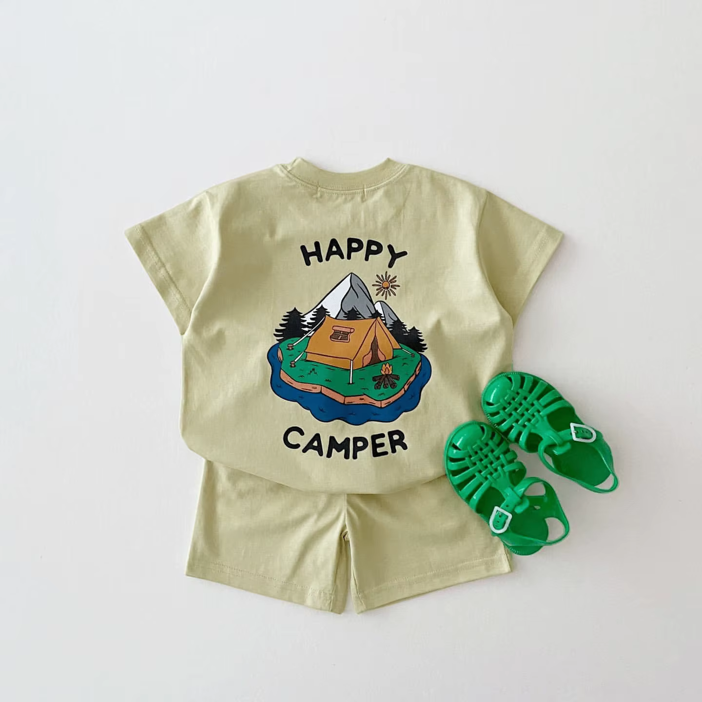 Komplekt 'Happy Camper' koosneb heledast T-särgist ja lühikestest pükstest, millel on värviline laagriteemaline trükk mägede, telgi ja päikesega. Taust on valge ja komplekti kõrval on rohelised sandaalid.