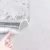 Käsi, mis koorib vana tapeedi alt Linane 3D tekstuuriga iseliimuvat paksu tapeeti valgel taustal, näidates realistlikku tekstuuri ja lihtsat paigaldust.