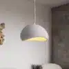 Skandinaavia stiilis LED laelamp helendab õrnas soojas valguses betoonseinalise ruumi taustal. Lamp on valge tekstuurse pinnaga ning ripub elegantselt minimalistliku disainiga interjööris.