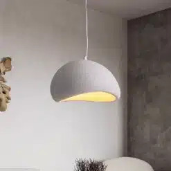 Skandinaavia stiilis LED laelamp helendab õrnas soojas valguses betoonseinalise ruumi taustal. Lamp on valge tekstuurse pinnaga ning ripub elegantselt minimalistliku disainiga interjööris.