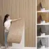 Naine paigaldab isekleepuvat paksu 3D elutoa seinatapeeti, millel on puiduvälimusega vertikaalsed ribad. Tapeet on heledas tooni ja kergesti pealepandav, luues sooja ja stiilse interjööri. Taustal on näha lihtne valge sein ja paremal valik erinevates toonides tapeeti.