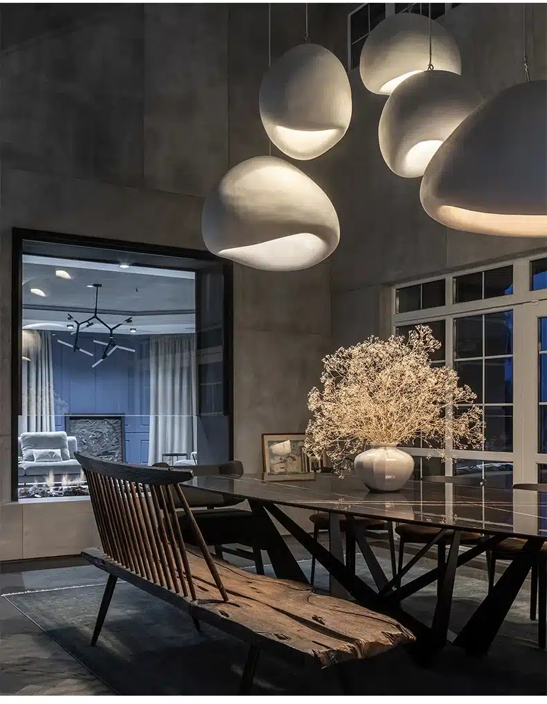 Elegantne Skandinaavia stiilis LED laelamp, mis ripub söögilaua kohal hämaralt valgustatud ruumis. Laelambid on valged ja stiilse kujuga, luues kaasaegse ja õhulise atmosfääri. Taustal on suur aken ning maja sisekujundus on maalähedane ja kaasaegne.
