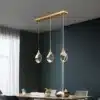 Kolm luksuslikku kristallidega ripplampi koos kuldsete riputustega rippumas elegantse söögilaua kohal, uhkeldades oma säravate LED-valgustitega tumedal taustal, Luksuslikud kristallidega ripplambid.