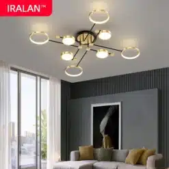 Kaasaegne elutuba, kus laes on luksuslik puldiga LED laelamp kuldsete ja mustade elementidega, särab soojas valguses. Hall tapeet ja hele diivan täiendavad interjööri avara aknaga taustal.