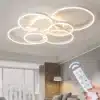 Stiilne LED laelamp, kujundatud mitme ringiga, loob hubase valguse ja kaasaegse atmosfääri magamistoas või elutoas. Kaugjuhtimispult käes võimaldab valguse juhtimist mugavalt. Taustal heledalt ja avaralt sisustatud ruum.