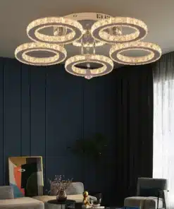 Luksuslik ruumis ripnev äpi või puldiga juhitav kristalllidega laelamp, mis valgustab elutuba soojas valguses. Lambil on viis elegantset rõngast, millel säravad kristallid, ja taustal on tume sinine sein.