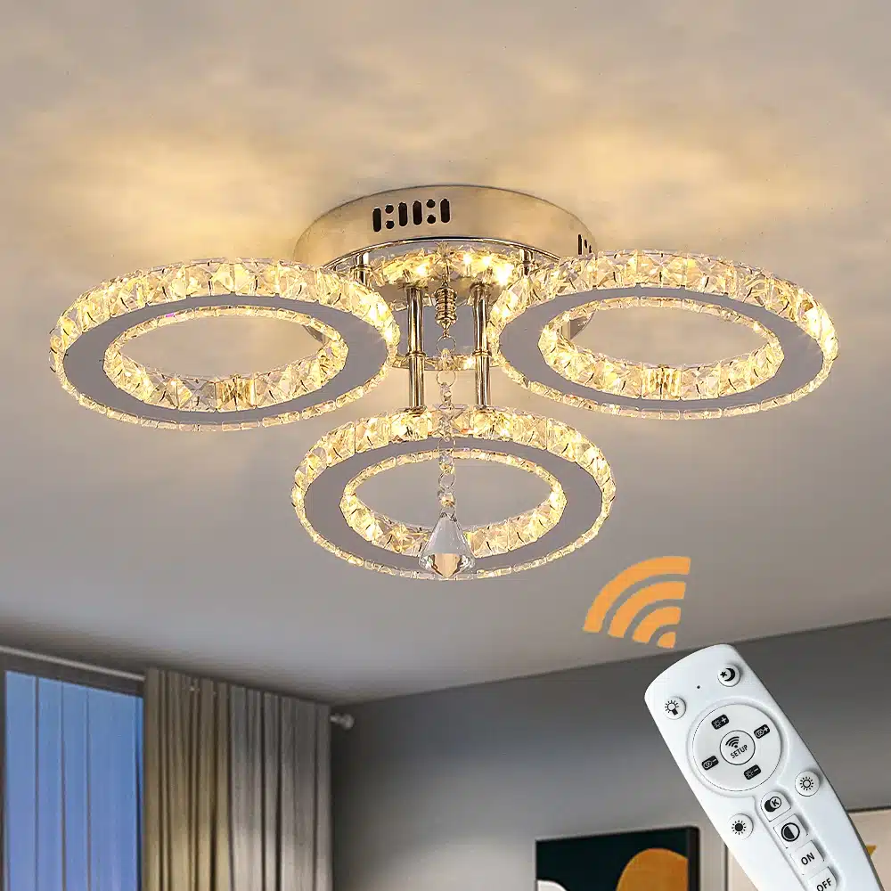 Kristallidega laelamp kolmest ringikujulisest LED-valgustist, mis ripub elegantses siseinterjööris. Lampi juhib kaugjuhtimispult, millel on erinevad valiknupud. Taustal hele sein ja kardinad.