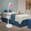 Võimas infrapuna füsioteraapia lamp valu leevendamiseks | E-pood Tootemaailm Ruumis seisab võimas infrapuna füsioteraapia lamp, valge korpuse ja reguleeritava varrega. Lamp kiirgab sooja punast infrapuna valgust, mis suunatud ühere uuele voodile taustal, sinise tekiga kaetud ning maitsekalt sisustatud.