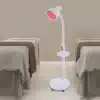 Võimas infrapuna füsioteraapia lamp valu leevendamiseks | E-pood Tootemaailm Modernne valge infrapuna füsioteraapia lamp punase valgusega, asetsev mugavas terviseruumis kahe voodi vahel beeži tausta ees.