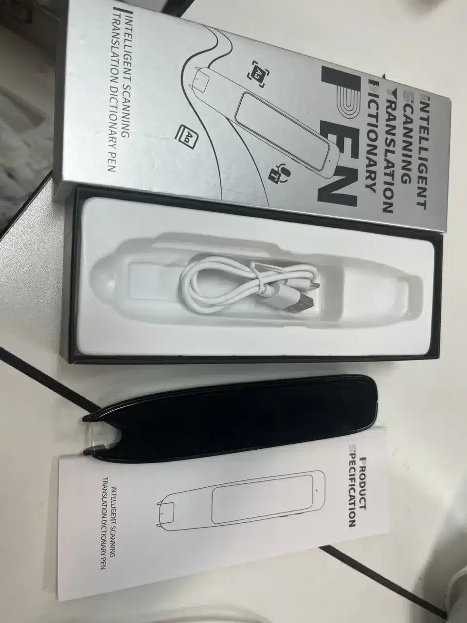 Nutikas tõlkepliiats - 142 keelt, hõbedases karbis koos laadimiskaabliga. Taustal kasutusjuhend ja must ümbris.