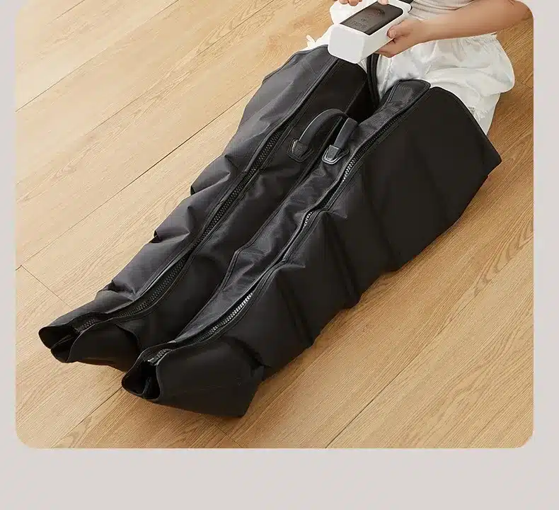 Isik kasutamas musta värvi õhusurvega massaažiseadet jalgadele veenilaiendite raviks, seadmel on juhtpult käes ning seade asub helepruunil parkettpõrandal.