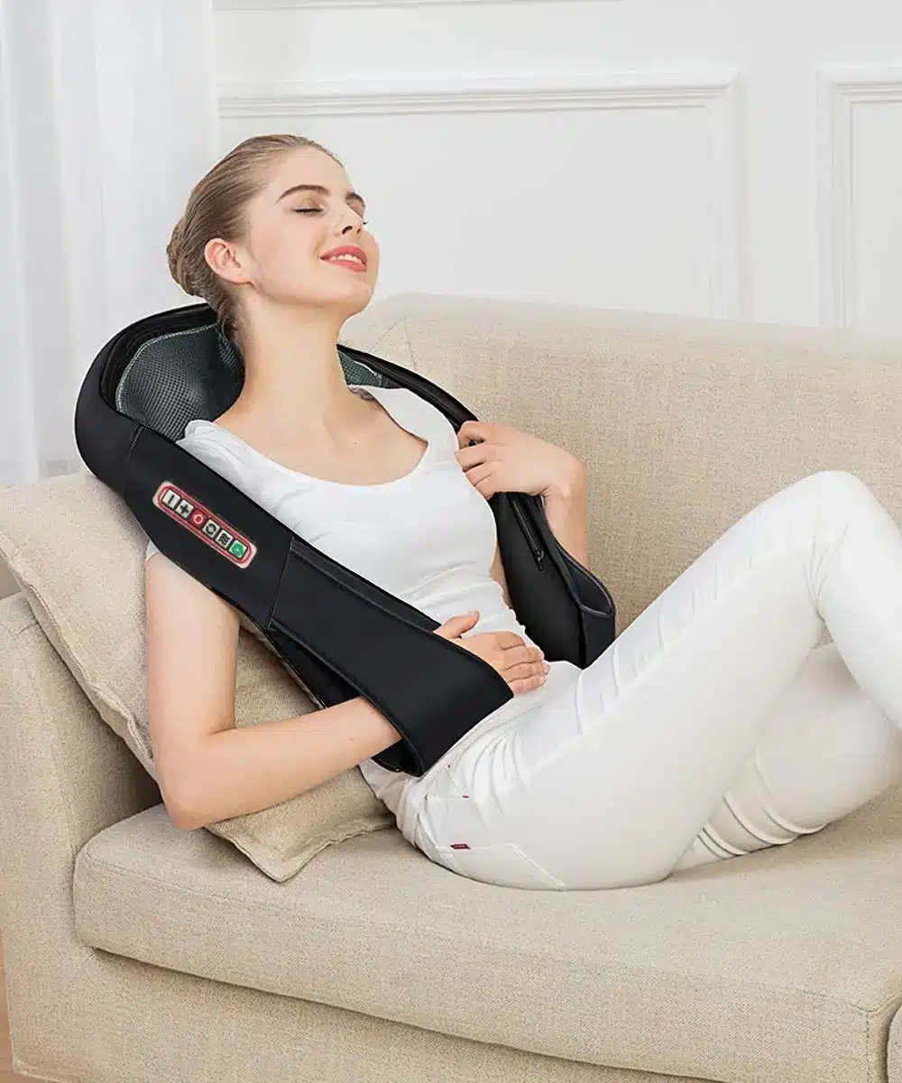 Naine istub diivanil ja naudib musta elektrilist õlamassažiaparaati, rahulolev ilme näol. Aparaat on valmistatud kvaliteetsest materjalist ja kohandub mugavalt õlgadele.