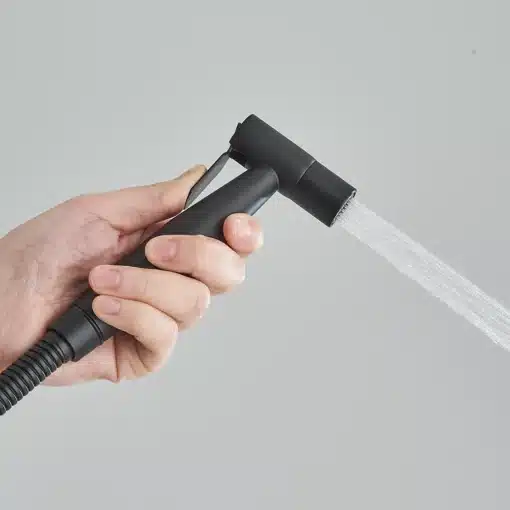 Seinale kinnitatav käimla bidet pihusti terasest | E-pood Tootemaailm Inimene hoiab hallis taustal käes musta terasest seinale kinnitatavat käimla bidet pihustit, millest voolab vett.