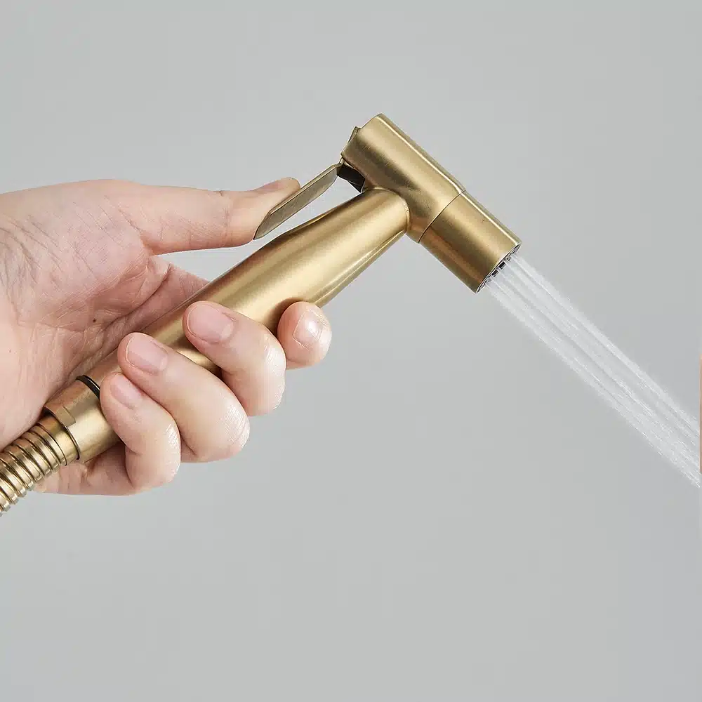 Kuldne terasest seinale kinnitatav käimla bidet pihusti, mida hoitakse käes ja millel on voolik ning veejuga, heleda taustaga.