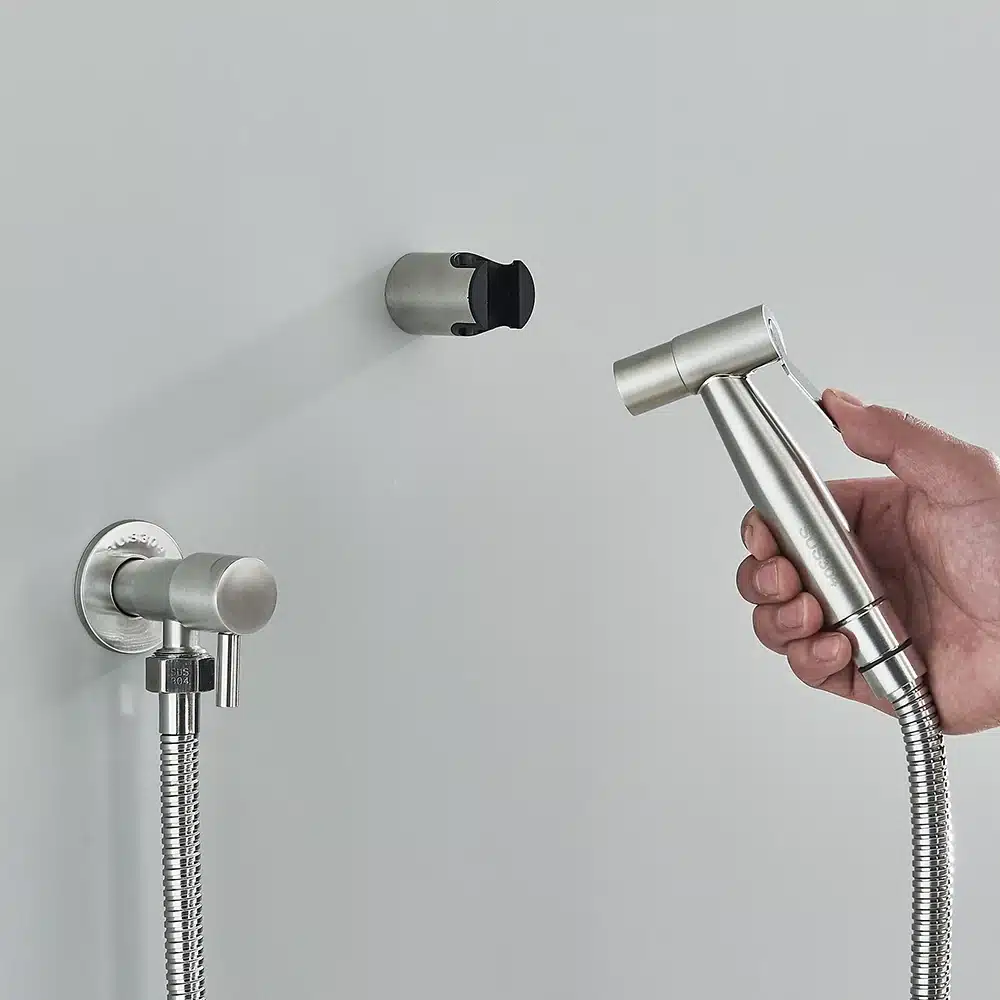 Hõbedane terasest seinale kinnitatav käimla bidet pihusti, mille käes hoiab käsi valge seina taustal. Pihusti voolik on paindlik ja metallist.