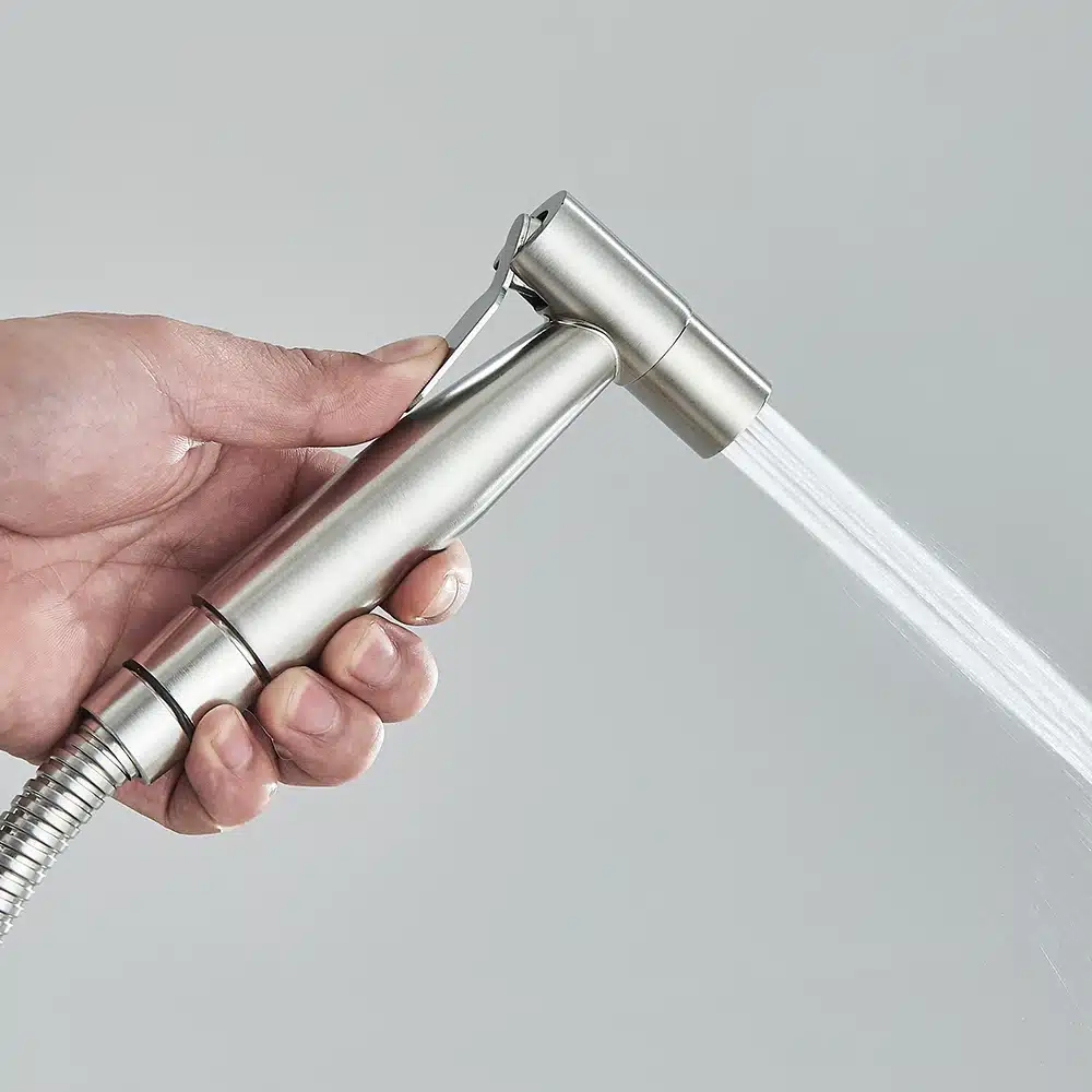 Käsi hoiab terasest seinale kinnitatava käimla bidet pihustit, kust voolab veejuga, halli tausta ees.